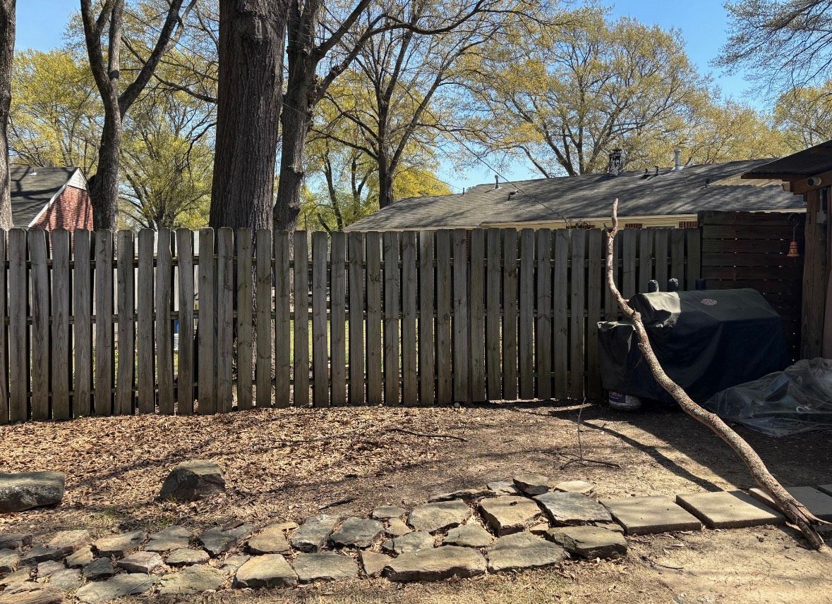 Wooden fence encloses a backyard with trees, a barbecue, and stone path under a sunny sky.