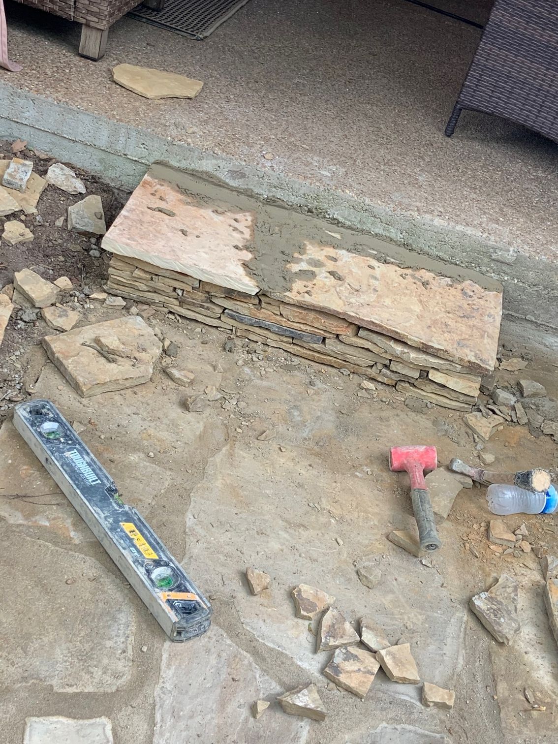 Stone patio being constructed; mortar, level, and hammer are visible.