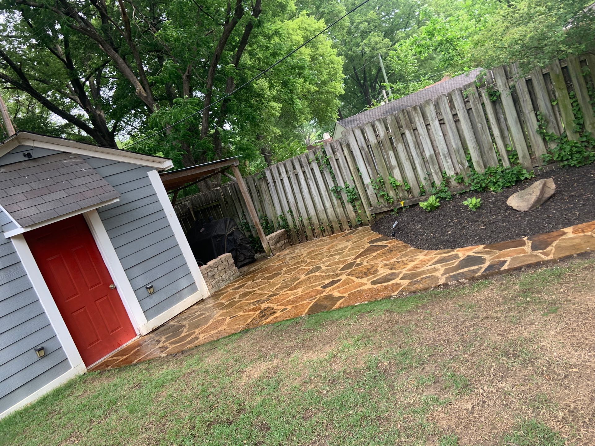Backyard with blue shed, red door, stone patio, wooden fence, and green grass.