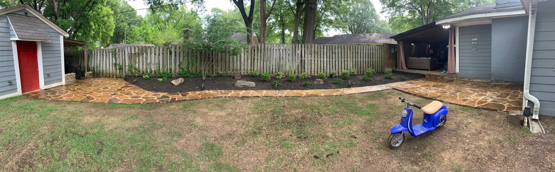 Backyard scene with sheds, a fence, mulch pathways, a blue scooter, and trees.
