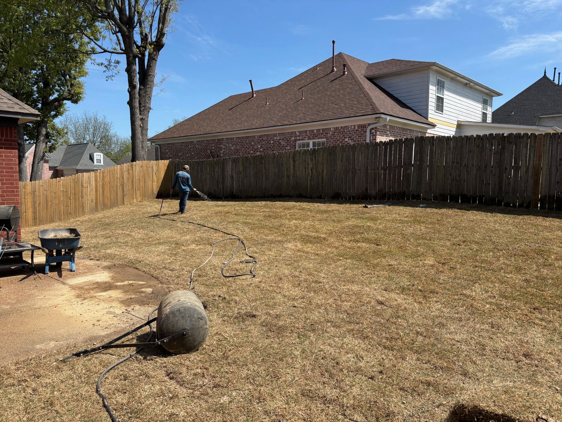 A person using a leaf blower in a backyard with dry grass, a wooden fence, and houses in the background.