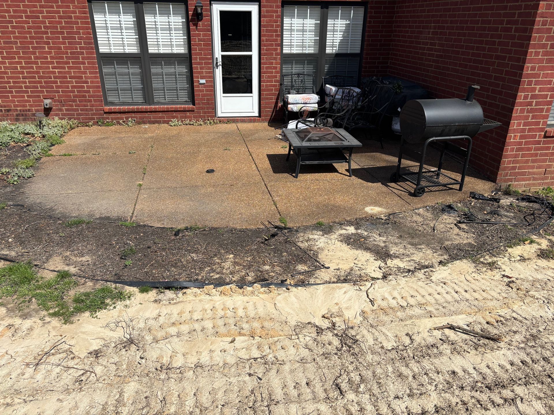 Brick patio with fire pit, grill, and door. The ground has sand, dirt, and tire tracks.