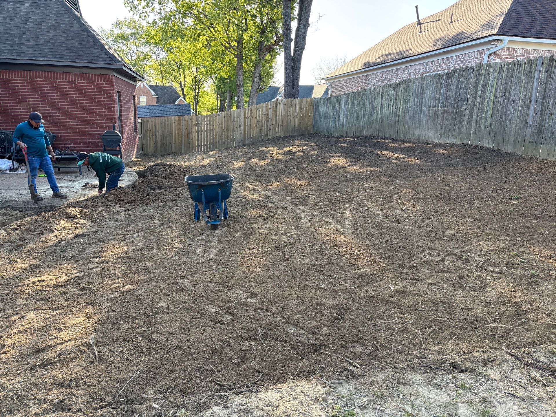 Two people preparing soil in a backyard near a brick building and wooden fence; one person uses a shovel.