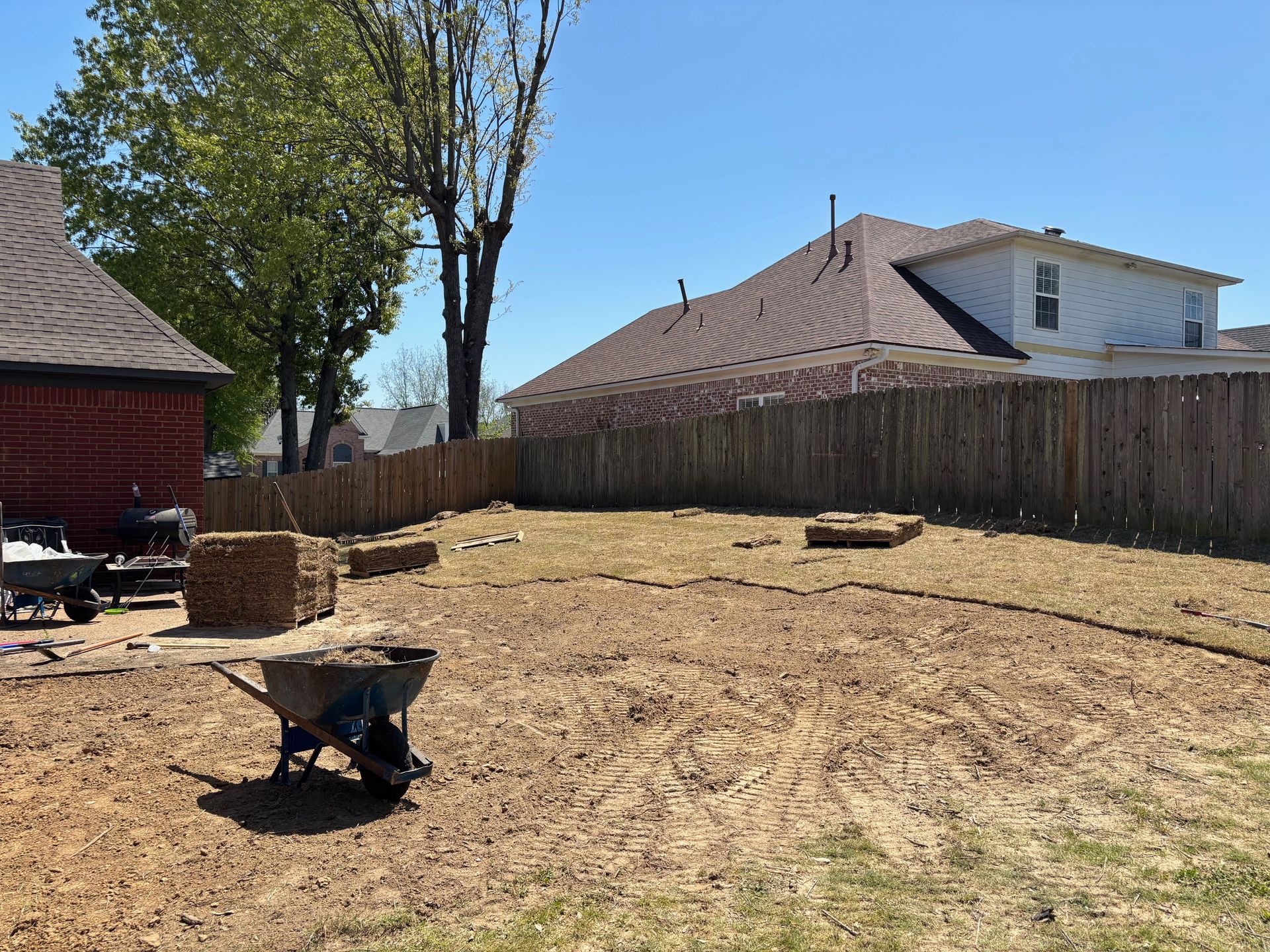 A mostly bare backyard with a wheelbarrow, hay bales, and a wooden fence under a bright blue sky.