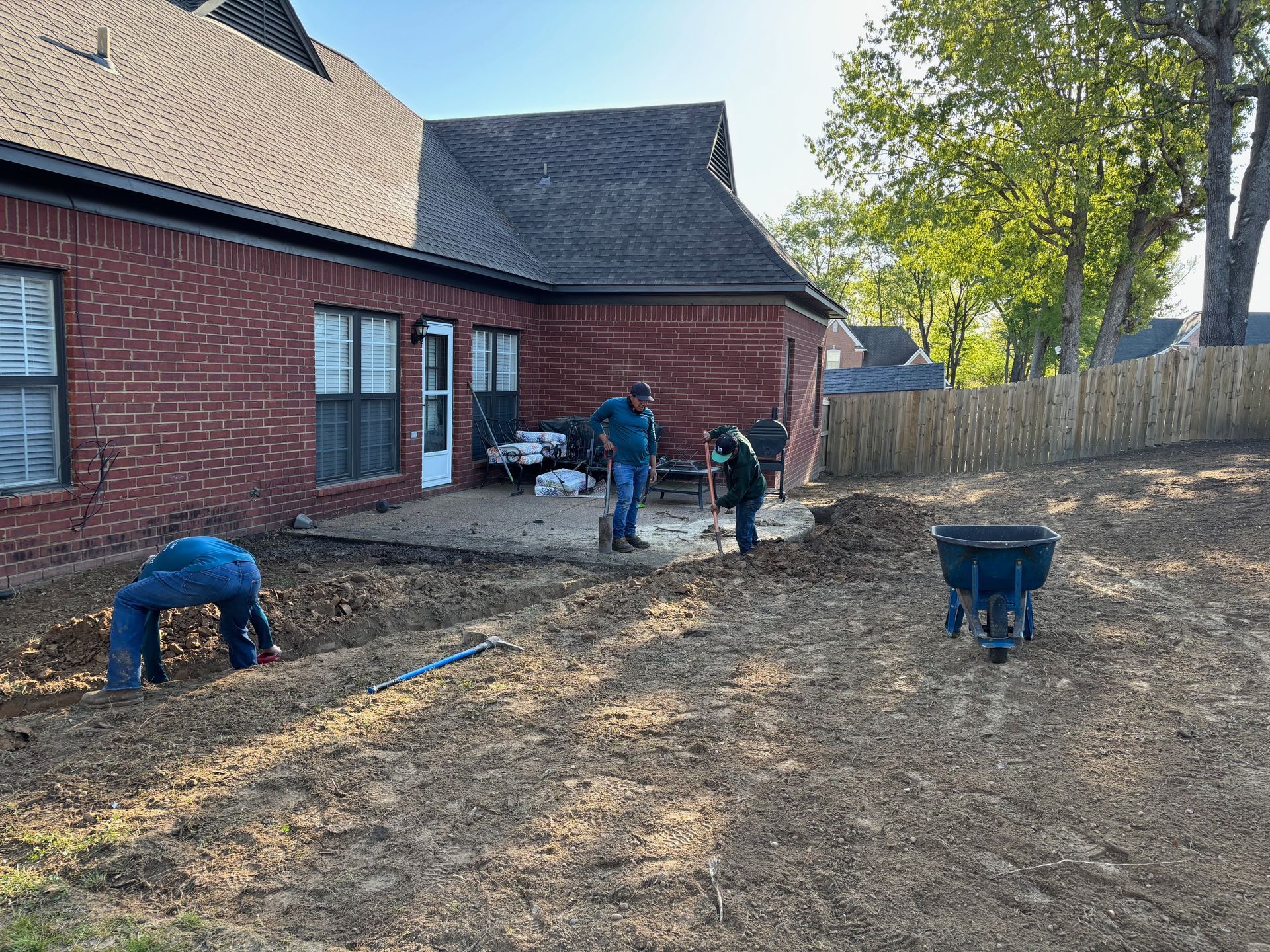 Three people working in a yard near a brick house. They are digging and preparing the ground for landscaping.
