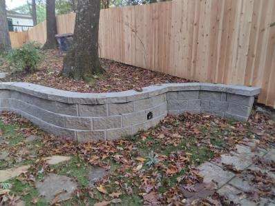 Gray block retaining wall curves around a tree, with a wooden fence in the background.