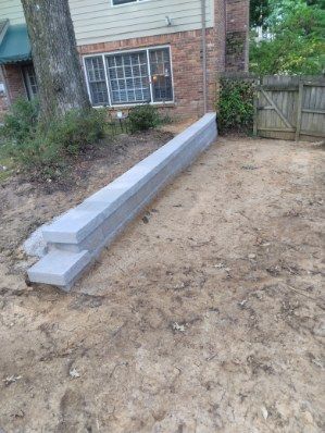 Gray concrete blocks stacked on dirt in front of a brick house, near a tree and fence.