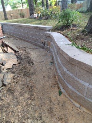 Curved retaining wall made of tan blocks, bordering a dirt path and yard.