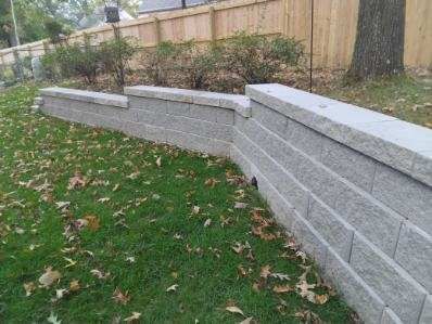 Gray retaining wall built on a slight slope in a yard with grass, leaves, and a wooden fence.