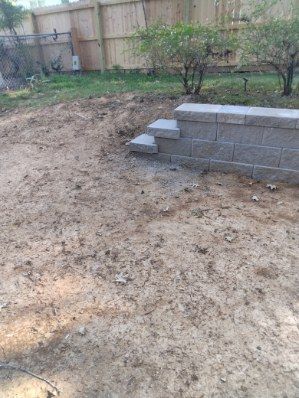 Dirt yard with a retaining wall and steps. Brown dirt with some grass and a wooden fence in the background.