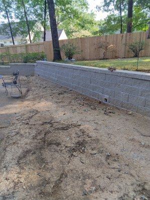 A gray block retaining wall borders a dirt patio. A wooden fence and trees are in the background.