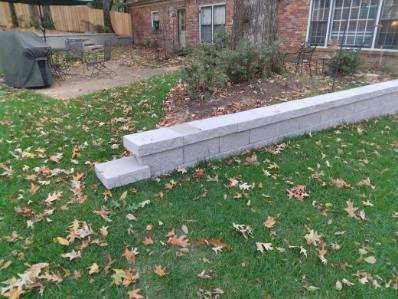 A concrete retaining wall with a step, on a grassy lawn with fallen leaves, in front of a brick house.