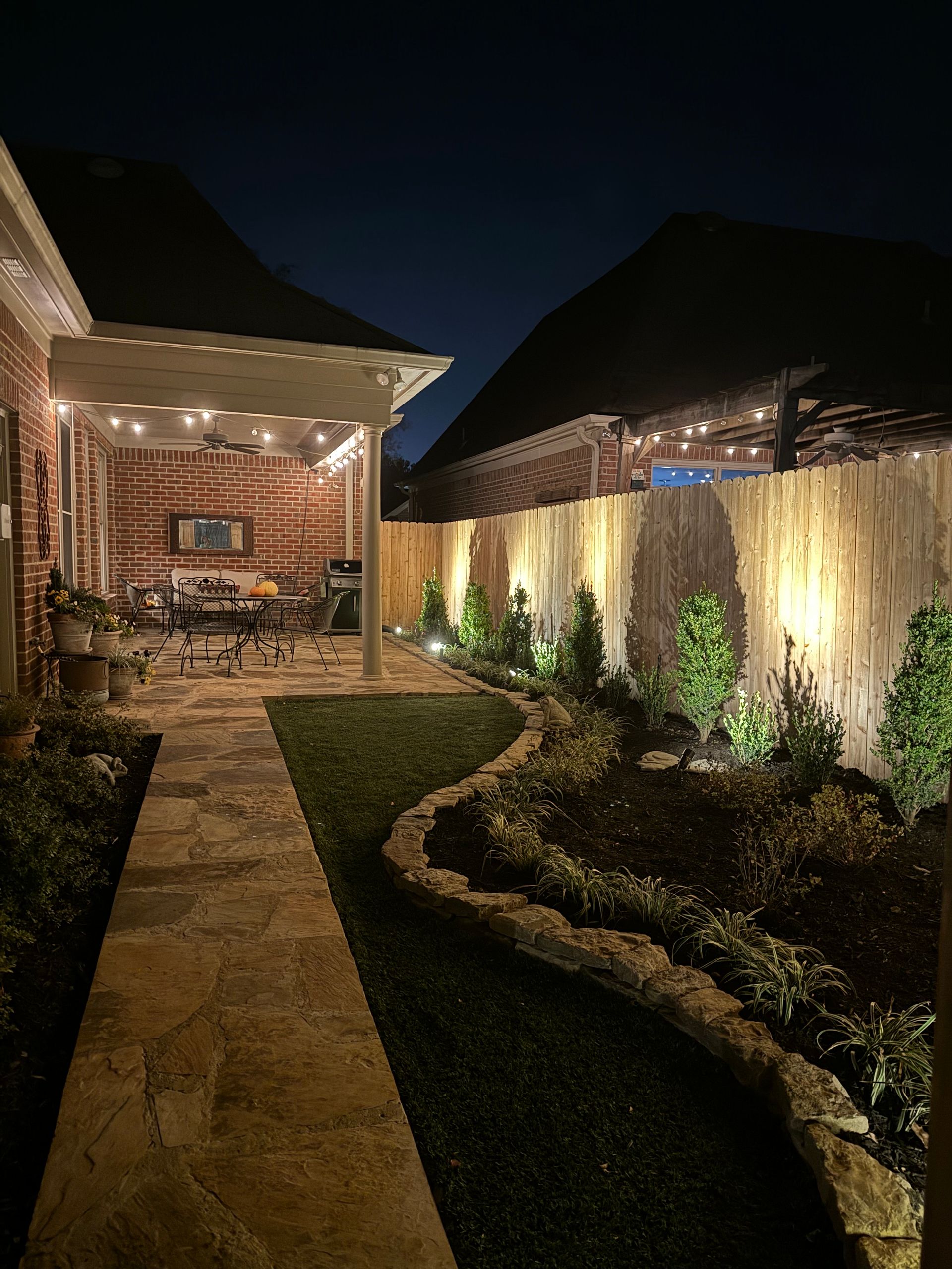 Nighttime backyard scene: illuminated patio, pathway, landscaping with lit bamboo fence.