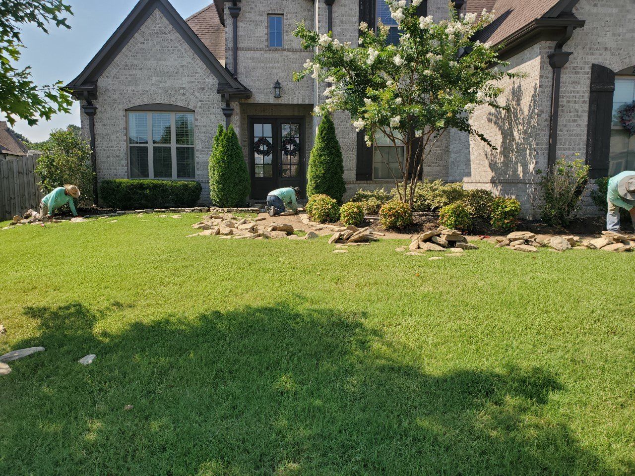 People working in front yard of a brick house, with manicured lawn and landscaping.