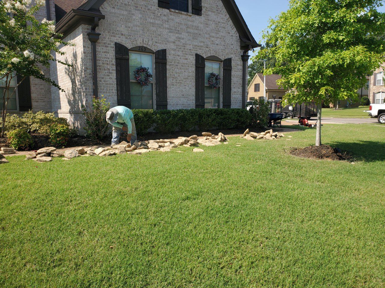 Person laying stones in front yard with green grass and a house.