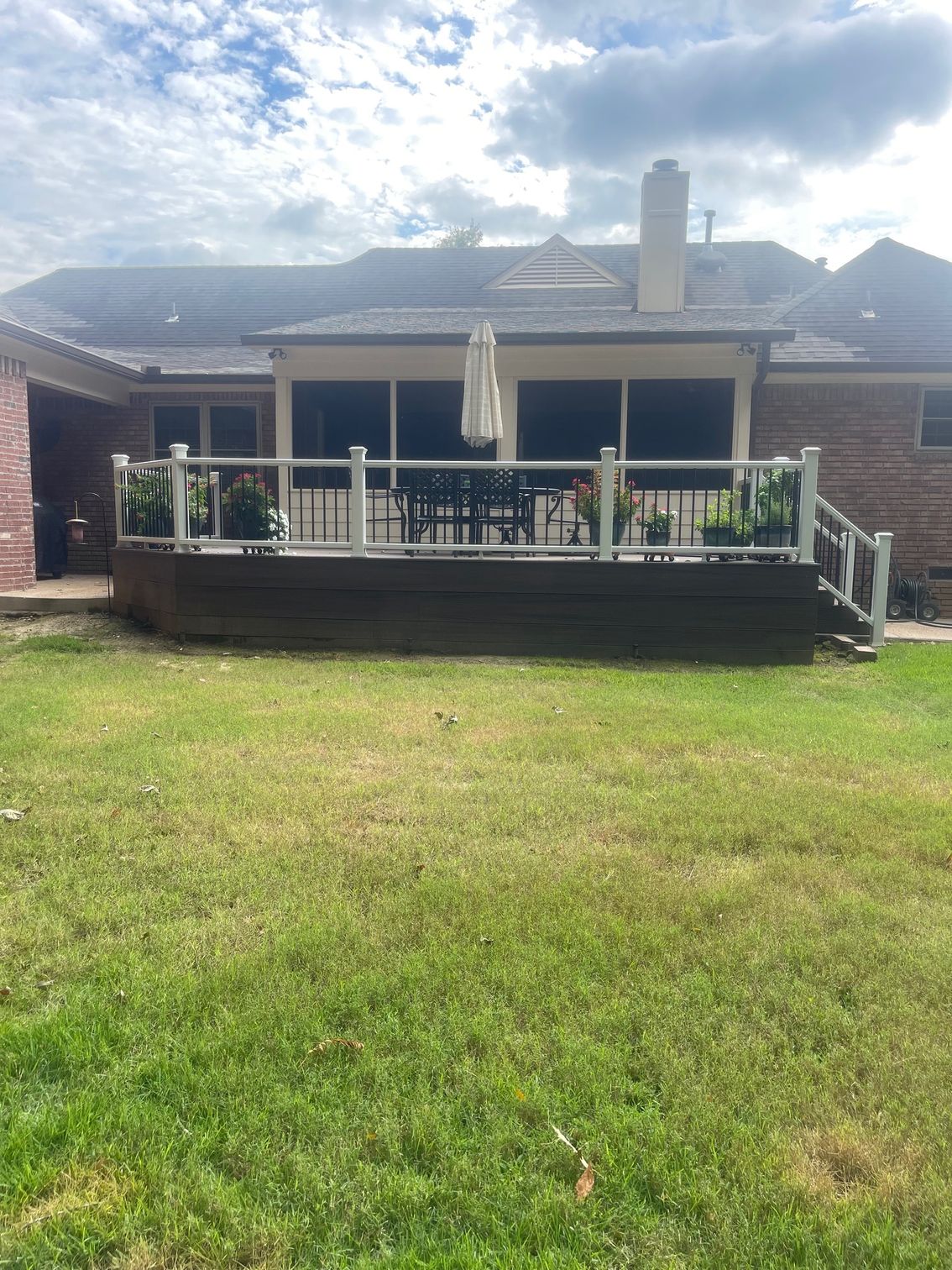 Backyard deck with white railing, table, chairs, and umbrella; green grass, brick house, cloudy sky.