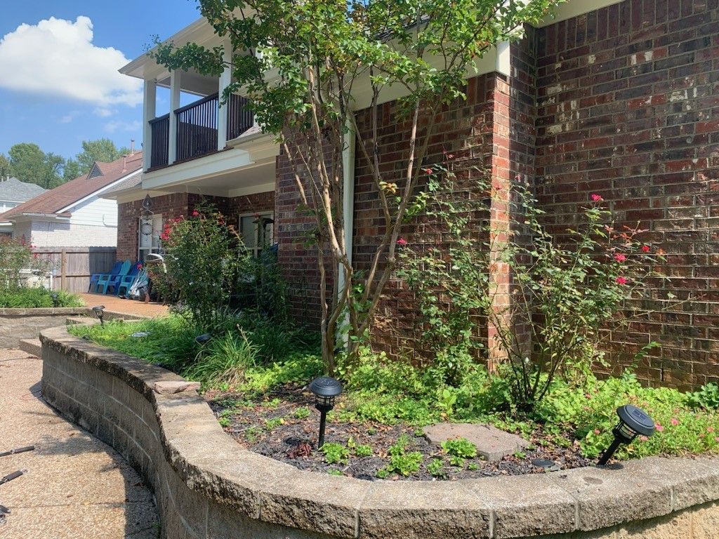 A brick building with a raised garden bed; walkway with solar lights; tree in the middle.