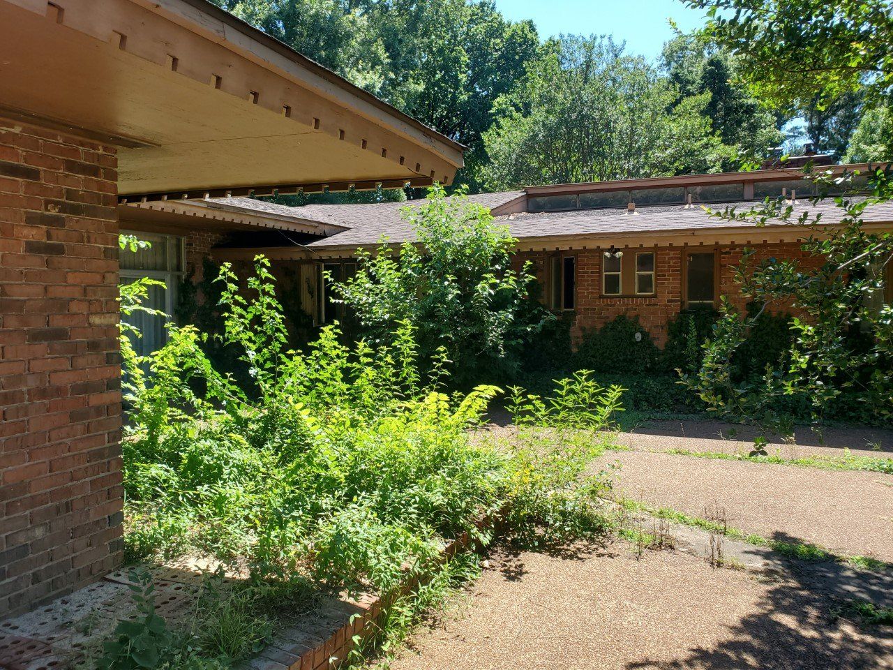 A brick building overgrown with green bushes and trees; a gravel driveway leads toward the entrance.
