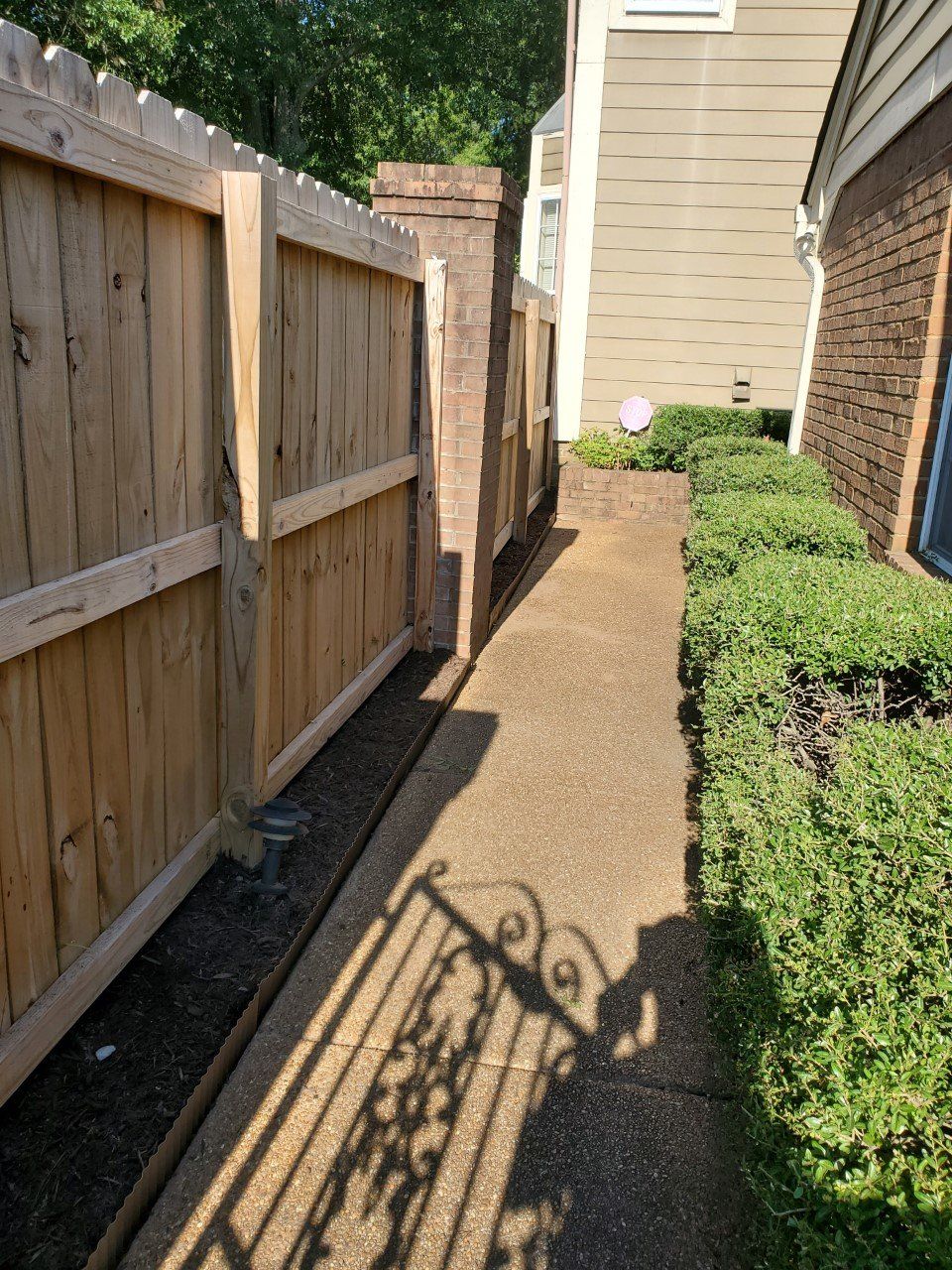 A narrow concrete walkway between a wooden fence and a brick building with trimmed bushes and a wrought iron shadow.
