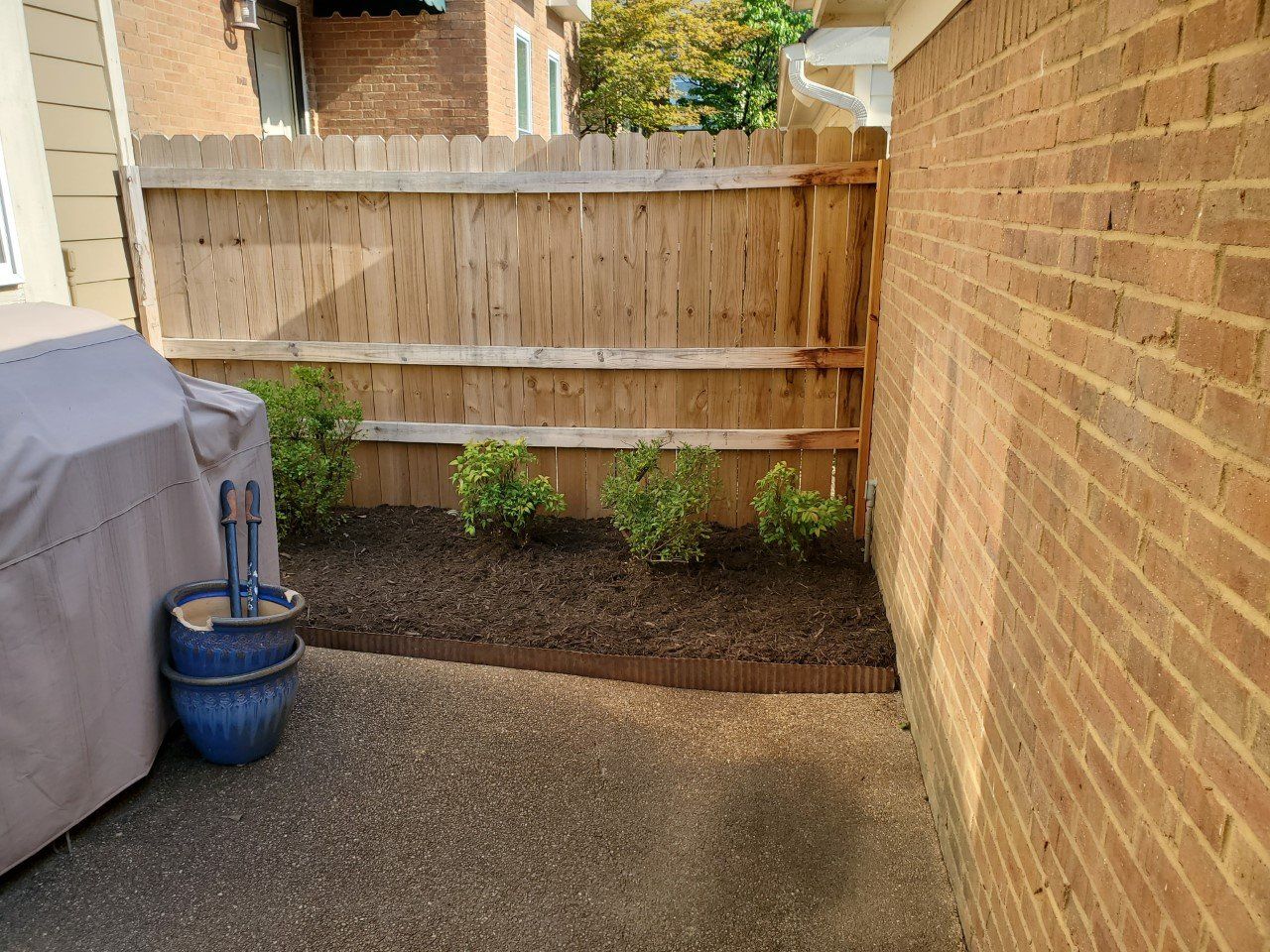 A small backyard patio with a wooden fence and brick wall. There is a garden bed and grill nearby.