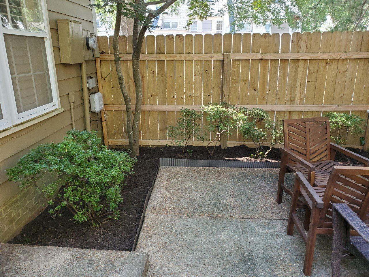 Small patio with wooden chairs, wooden fence, and bushes.