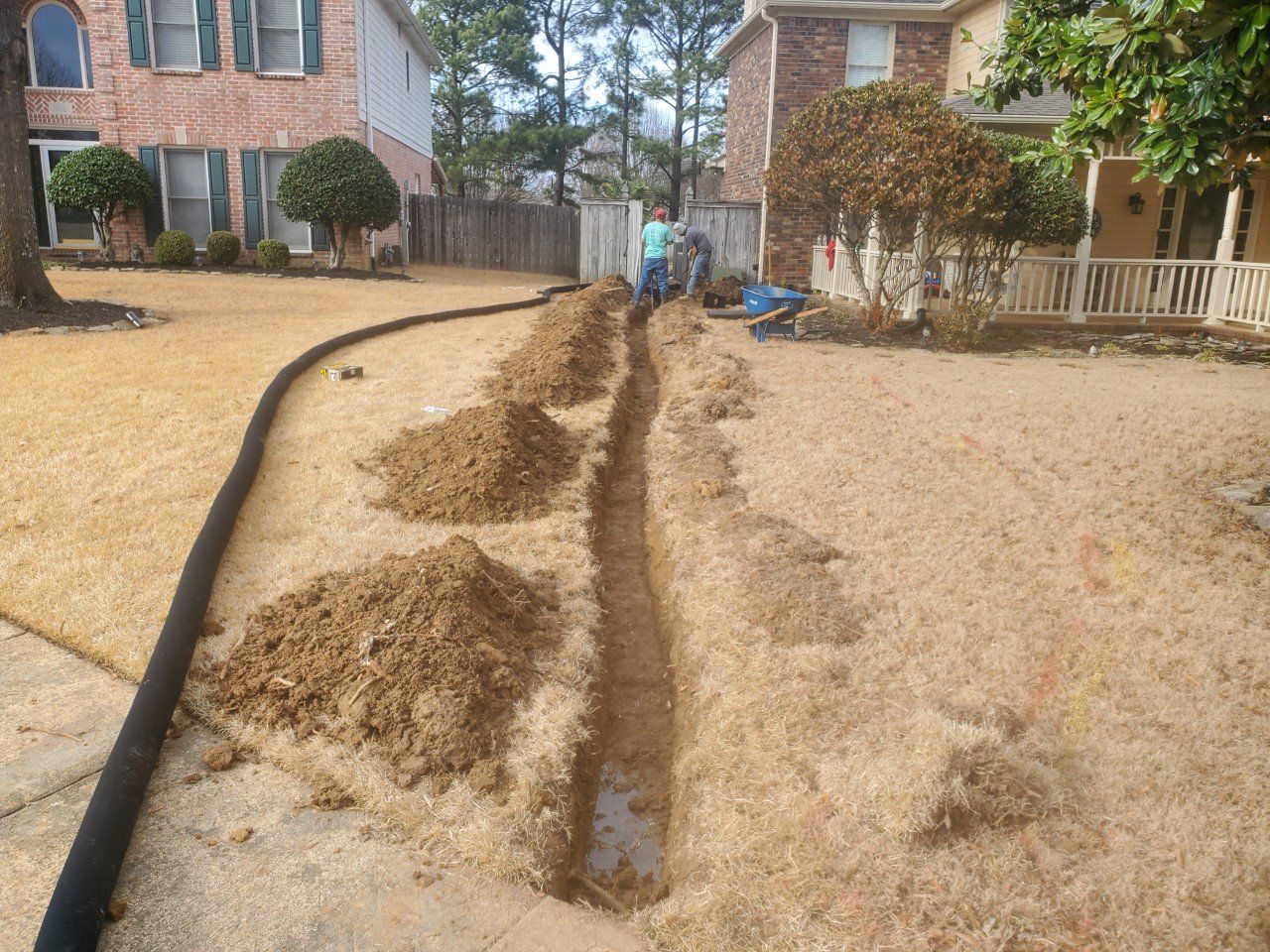 Workers digging a trench in a yard; soil piles beside it. A black tube runs along the grass.