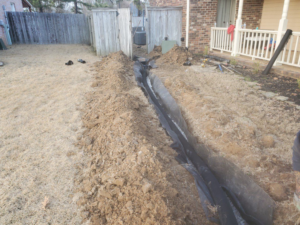 Trench dug in yard, lined with black material. Dirt mounds alongside a wooden fence and a house.