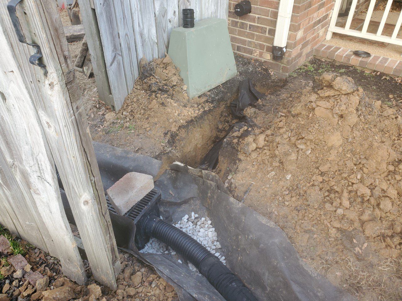 A trench with a black pipe near a fence and house, showing a drainage system being installed.