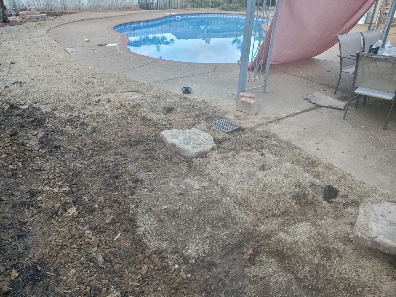 Backyard with a pool, concrete patio, and dirt/mulch. A pink shade covers a table with chairs.