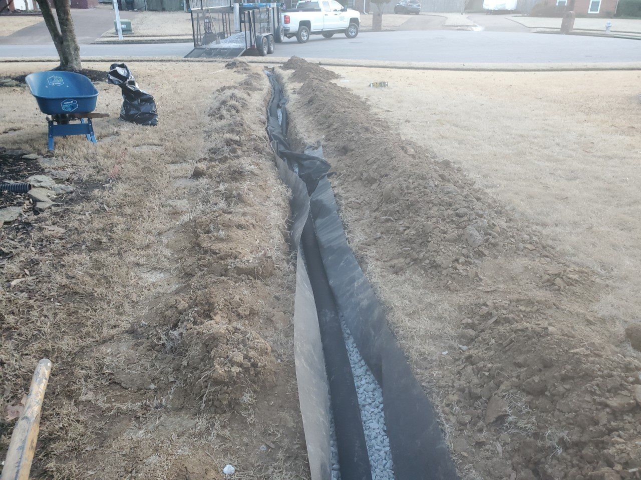 A trench dug in a grassy lawn with gravel at the bottom, equipment in the background.