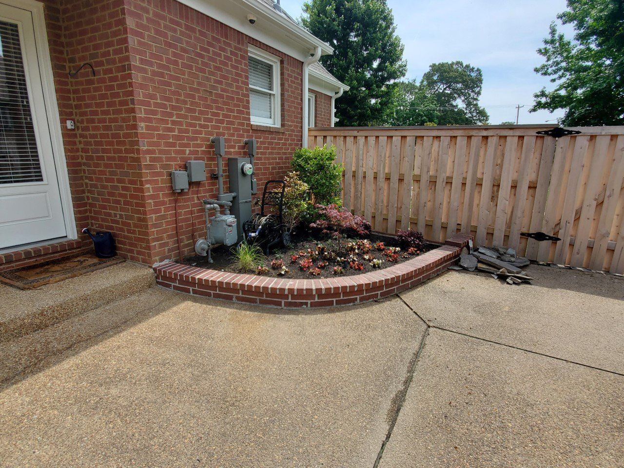 Brick house corner with a curved flower bed, brown fence, and concrete patio.