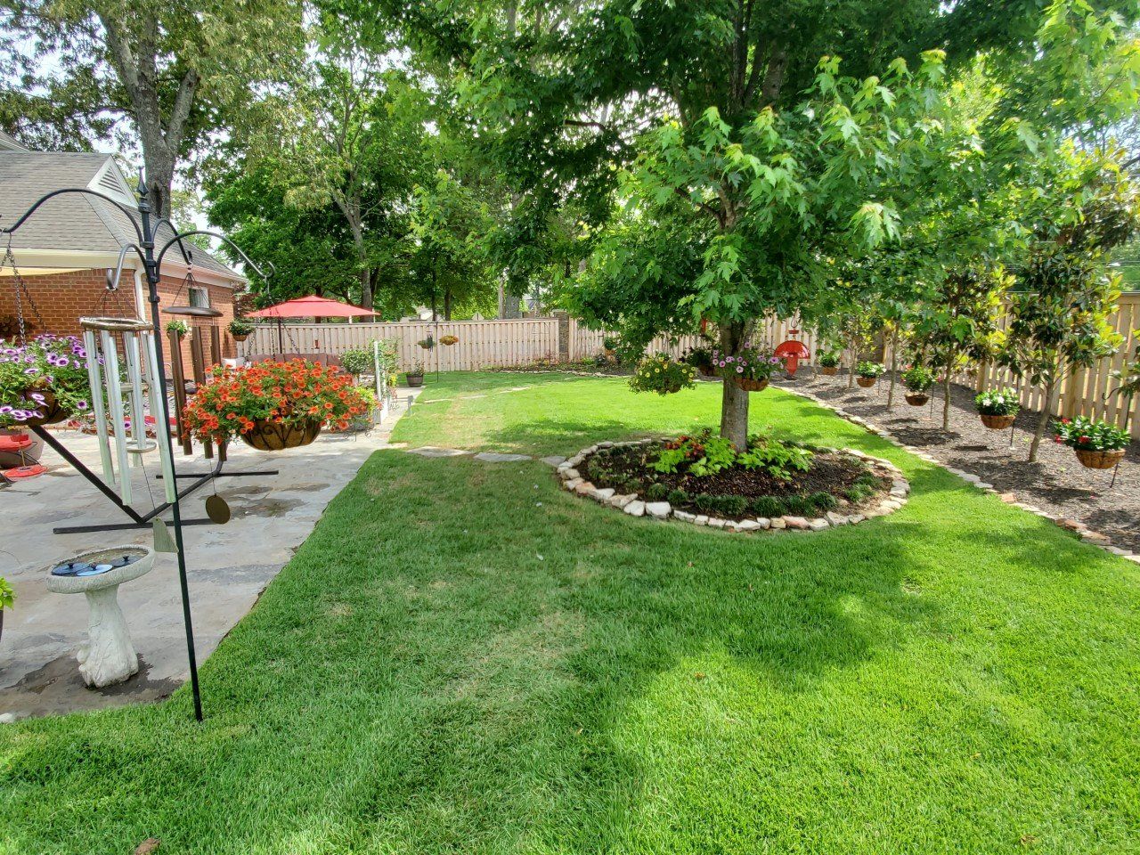 Green backyard with lush grass, a tree in the center surrounded by mulch and flowers.