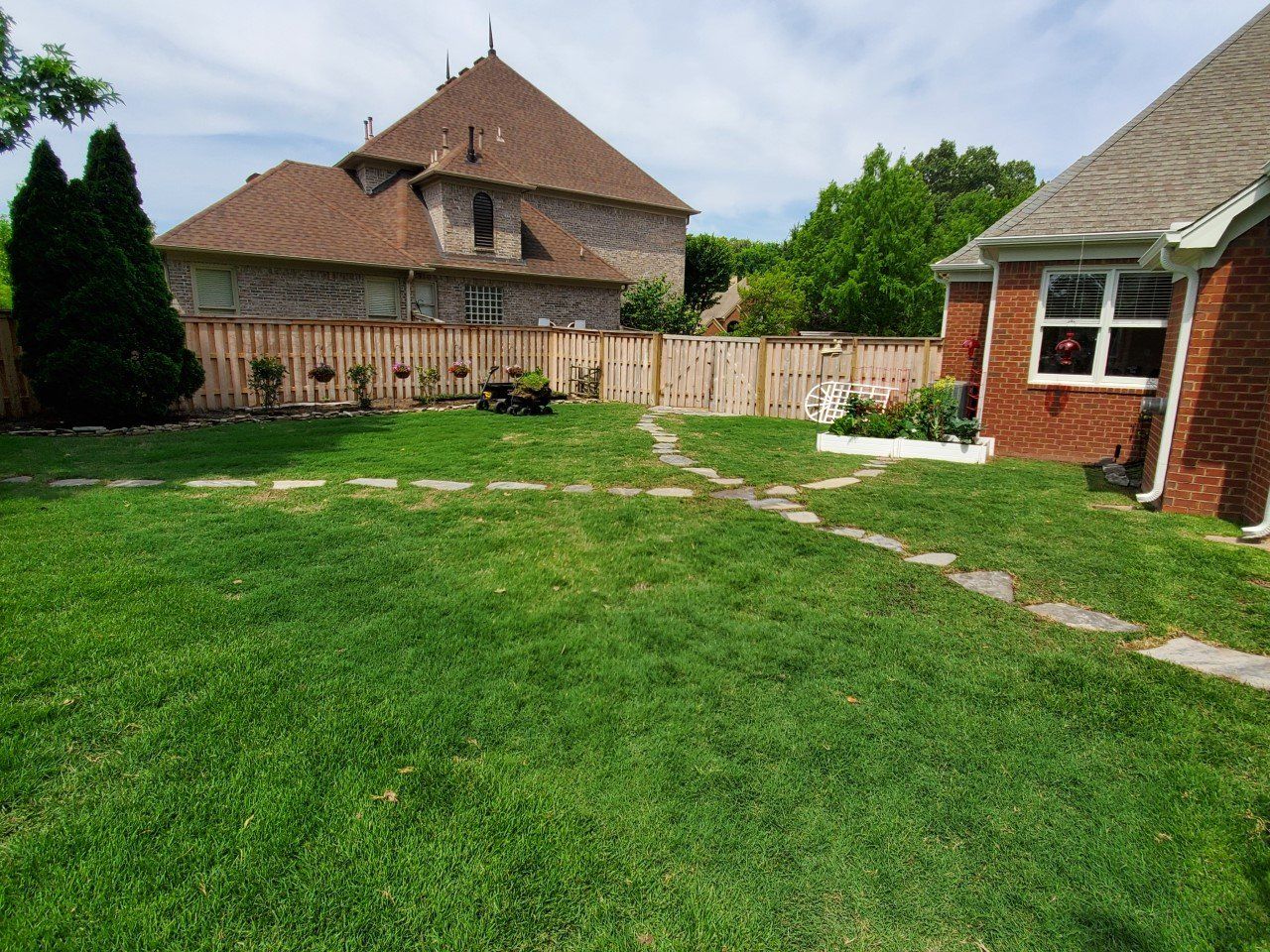 Green backyard with stone path, wooden fence, brick house, and a larger house in the background on a sunny day.