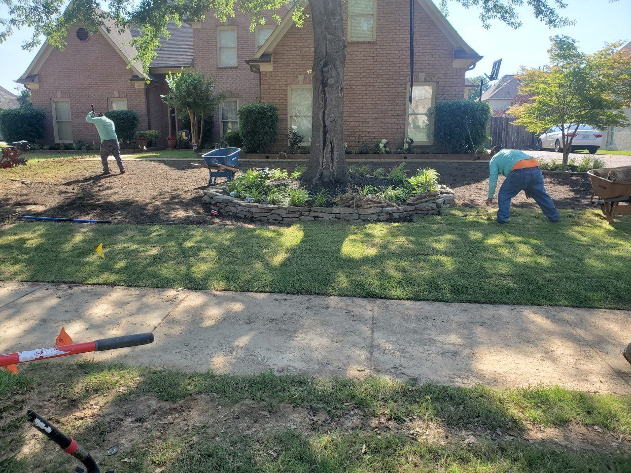 Two people landscaping a front yard with a large house in the background. Sunny day, green grass.