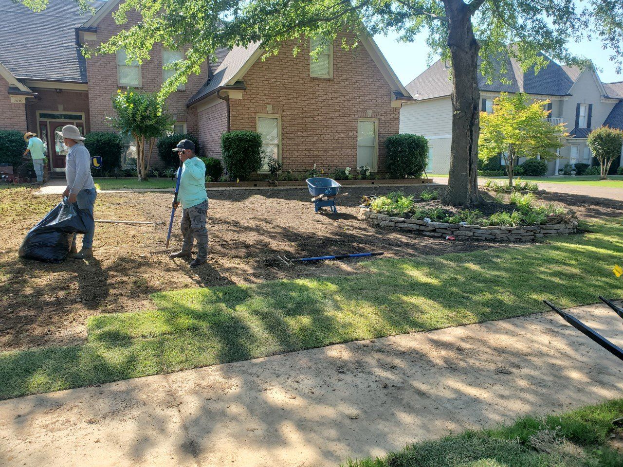 Landscapers working on a lawn near a brick house. One holds a bag, another rakes dirt.