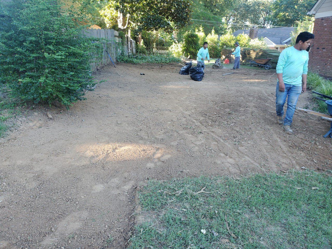 Men leveling dirt in a yard, preparing for landscaping. Green grass, foliage and a house are nearby.