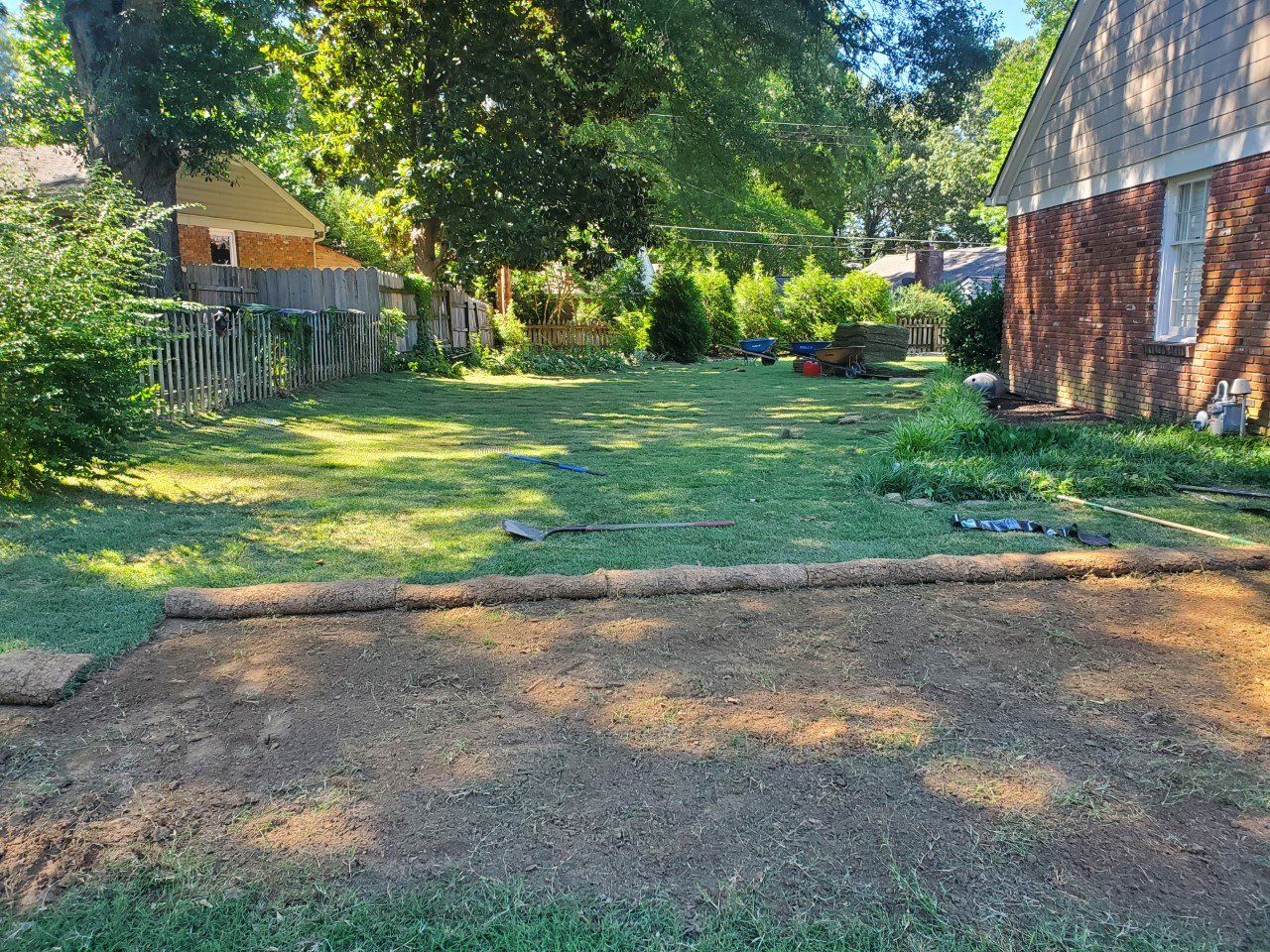 Backyard with newly laid soil in foreground, grass, trees, fence, and houses.