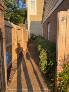 Narrow walkway between a wooden fence and building; sunlight casts long shadows.