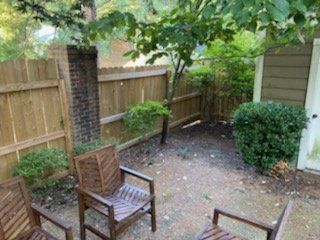 Patio with wooden chairs, a small tree, fence, brick pillar, and green shrubs.