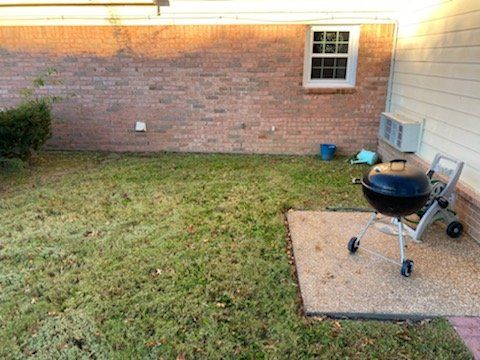 A backyard with a grill on a patio, a brick wall, window, and grassy area.