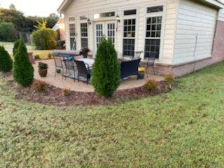Backyard patio with table, chairs, green shrubs, and lawn. Light-colored siding on the house.