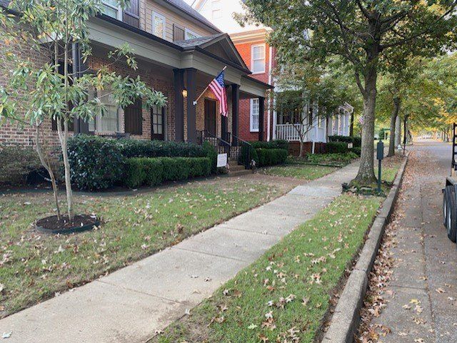 Sidewalk in front of brick houses with green lawns and trees on a sunny day. An American flag is visible.