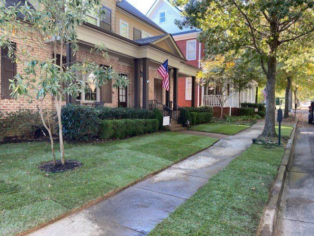 Sidewalk with fresh green grass and trees next to a brick house with an American flag.