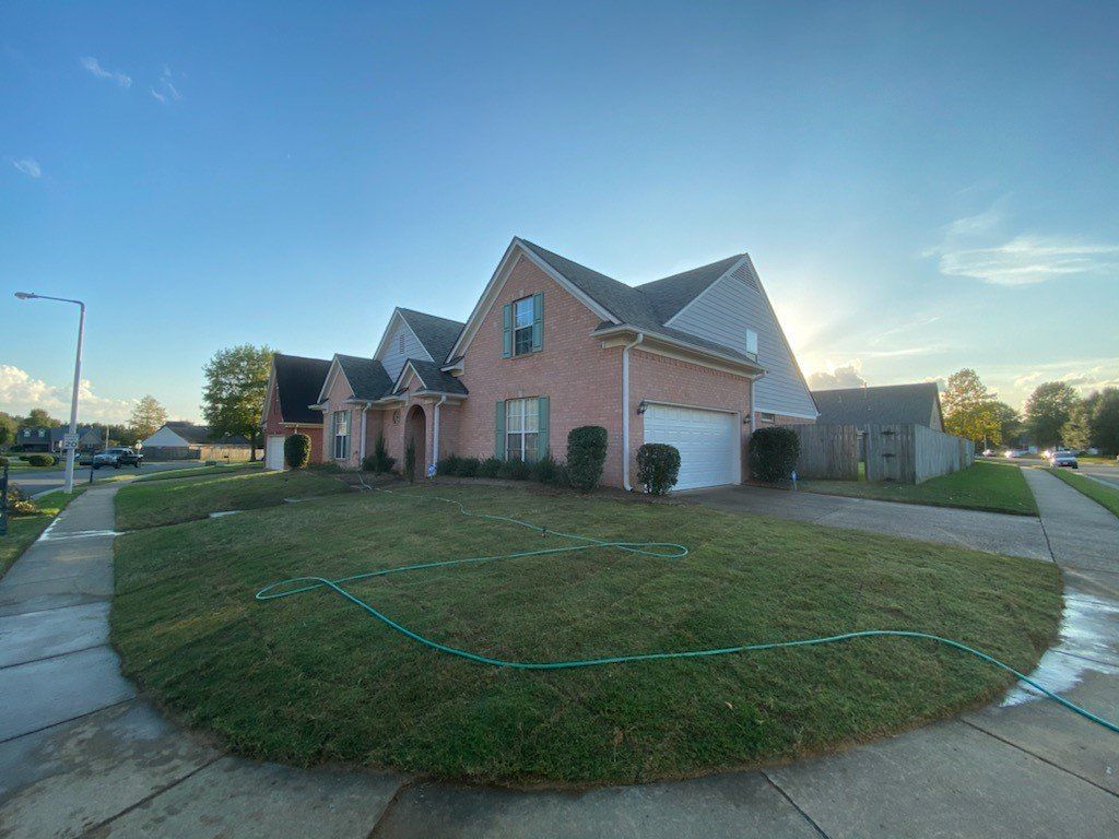 A suburban home with brick facade and lush green lawn under a blue sky. A green hose is visible.