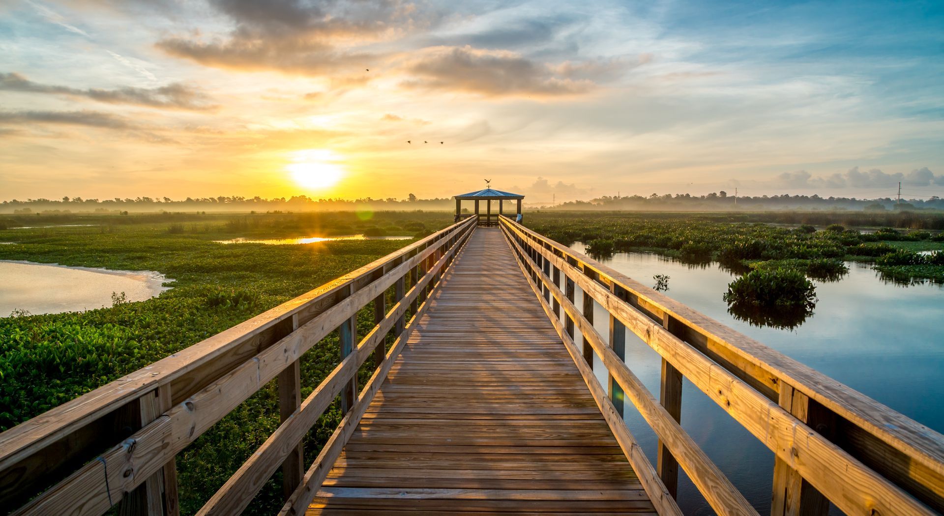 A wooden walkway leading to a gazebo overlooking a swamp at sunset.