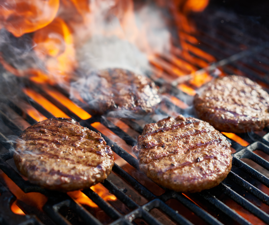Three hamburger patties are cooking on a grill with flames behind them