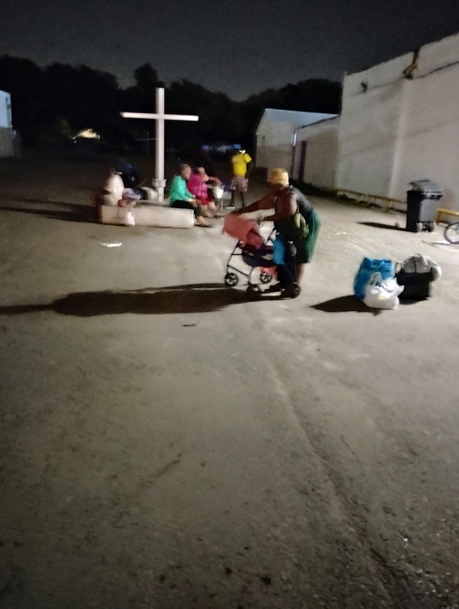 A group of people are gathered in a parking lot at night with a cross in the background.