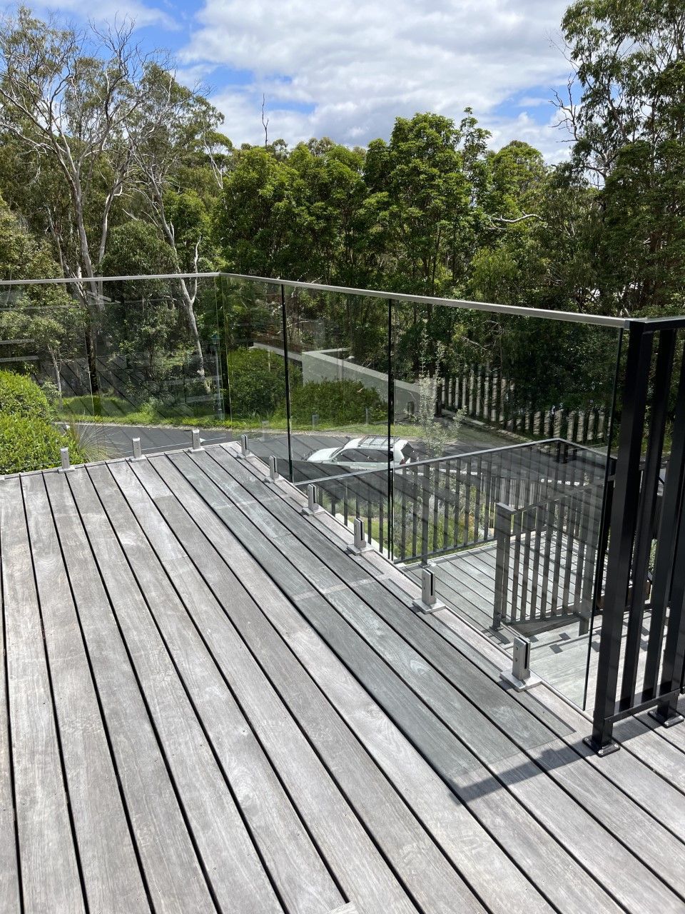 A wooden deck with a glass railing and trees in the background — Fabricator In Coffs Harbour, NSW