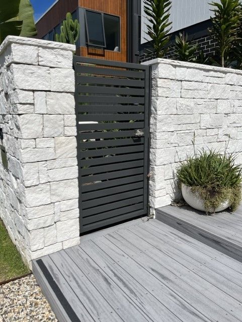 A stone wall with a black gate leading to a house. — Fabricator In Coffs Harbour, NSW