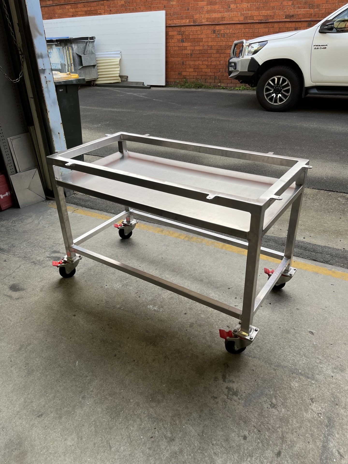 A stainless steel cart with wheels is sitting on the ground next to a car — Fabricator In Coffs Harbour, NSW